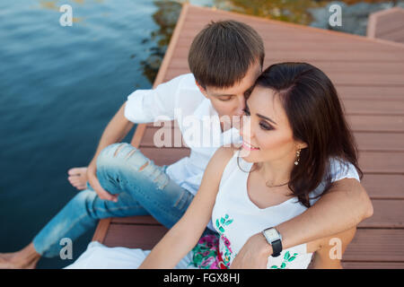 man embraces girl sitting on a pier at the river bank Stock Photo