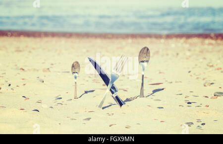 Vintage toned crossed knife and fork stuck in sand, shallow depth of field. Stock Photo