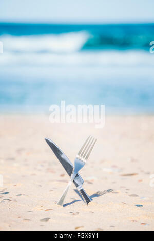 Crossed processed knife and fork stuck in sand, shallow depth of field. Stock Photo