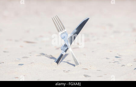 Crossed knife and fork stuck in sand, shallow depth of field. Stock Photo