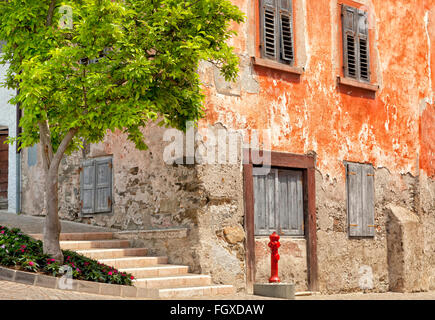 Old red historic corner house with aged walls and shutter windows, on cobbled street, with red water hydrant and tree in front Stock Photo