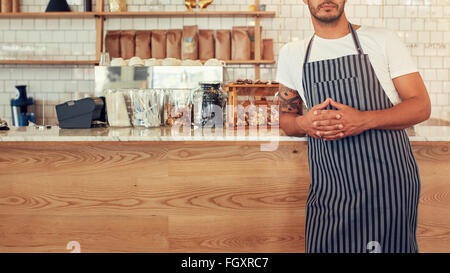 Cropped shot young man standing at a coffee shop counter wearing an apron. He is leaning to the cafe counter with his hands clas Stock Photo