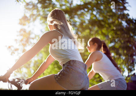 Two young women girlfriends wearing jeans shorts riding bicycles  in park on sunny summer day, back view Stock Photo