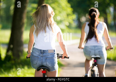 Two young beautiful women girlfriends wearing jeans shorts biking on sidewalk on the street on sunny summer day, back view Stock Photo