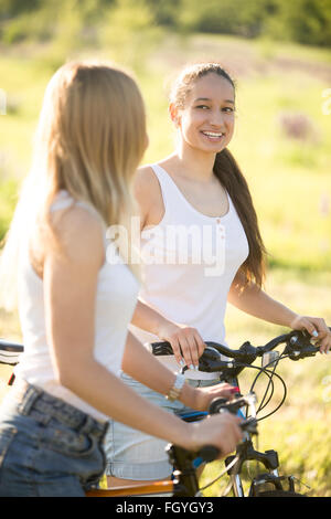 Two young beautiful cheerful women girlfriends wearing jeans shorts on bicycles in park on sunny summer day, having good time Stock Photo