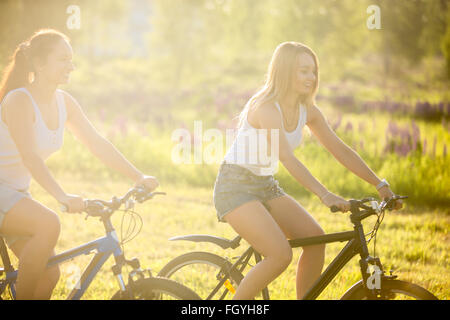 Two cute young happy smiling beautiful women girlfriends wearing jeans shorts riding bikes in park in bright sunlight on summer Stock Photo