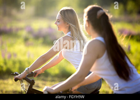 Two cute young happy smiling beautiful girlfriends wearing casual white tank tops enjoy cycling in park in bright sunlight Stock Photo
