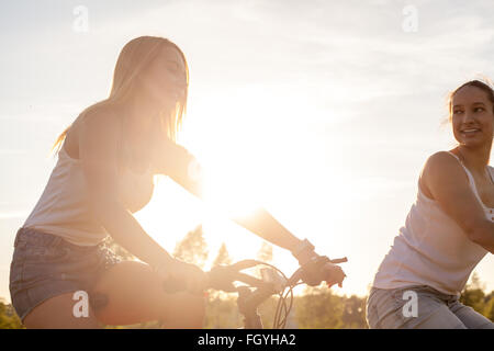 Two pretty young happy smiling beautiful girlfriends wearing casual white tank tops and jeans shorts riding bikes in park Stock Photo