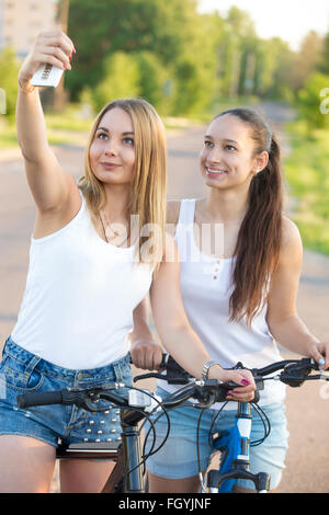 Two happy smiling beautiful young women friends wearing casual clothes standing with bikes taking self portrait on the road Stock Photo