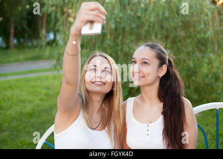 Two happy smiling beautiful young women friends wearing casual clothes sitting on park bench on summer day taking selfie Stock Photo