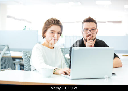 Two thoughtful young business people sitting in the office and using laptop together Stock Photo
