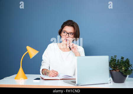 Thoughtful beautiful young woman in glasses sitting at the table and writing in notebook over blue background Stock Photo