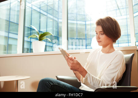 Pretty pensive young woman sitting in office and using tablet Stock Photo