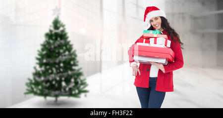 Composite image of festive brunette in santa hat and red coat holding pile of gifts Stock Photo