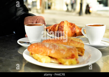 man sitting in a table in the terrace of a cafe set for breakfast, with some cups of coffee, a croissant and a spanish omelette Stock Photo