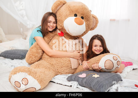 Two happy lovely sisters sitting and hugging plush bear in playroom Stock Photo
