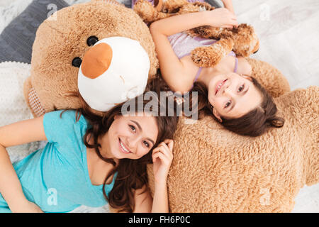 Top view of two smiling beautiful sisters lying on big soft plush bear in children room Stock Photo