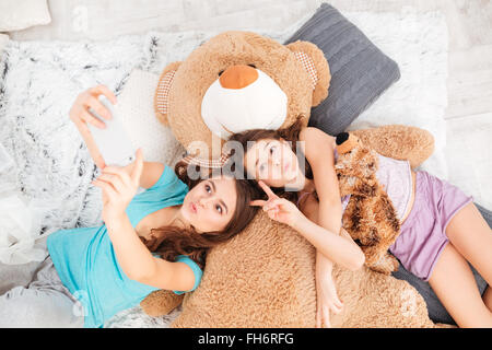 Two playful cute sisters lying, sending kisses and making selfie using smartphone at home Stock Photo