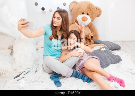 Two charming happy sisters making duck faces and taking selfie in children room Stock Photo