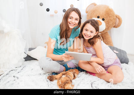 Two charming smiling sisters sitting and using tablet together in children room Stock Photo