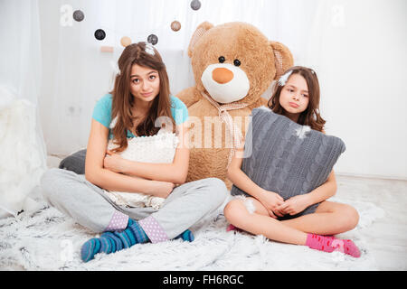 Two sad tired sisters sitting and hugging pillows in children room at home Stock Photo
