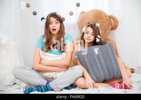 Two amazed lovely sisters sitting and hugging pillows at home Stock Photo