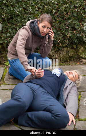 senior adult is lying on the ground, young woman makes an emergency call Stock Photo