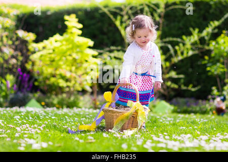 Little girl playing in sunny garden. Baby on Easter egg hunt in flower meadow. Toddler child with decorated basket picking eggs Stock Photo