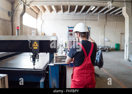 view from behind of a man wearing red overalls and a white construction helmet, standing, operating the control panel of a robot Stock Photo
