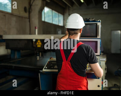 view from behind of a man wearing red overalls and a white protective helmet, standing, operating the control panel of an indust Stock Photo