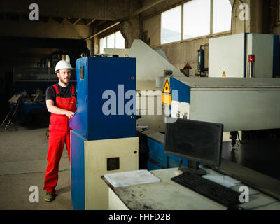 view from the right side of a worker looking busy, wearing red overalls and a white protective helmet, standing, operating the c Stock Photo