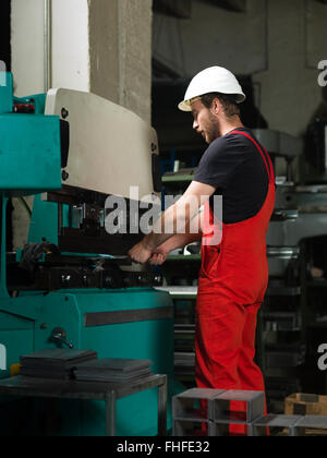 left side view of a worker, standing, wearing red overalls, and a white protective helmet operating an industrial machinery, pai Stock Photo