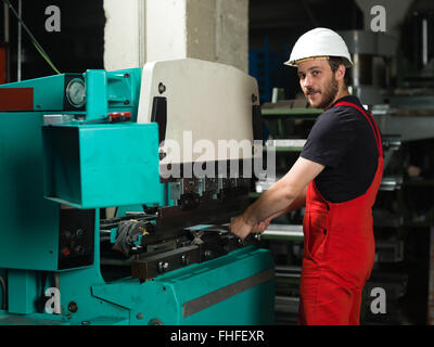 left side view of a worker, standing, wearing red overalls, and a white protective helmet holding a metal plate inside an indust Stock Photo