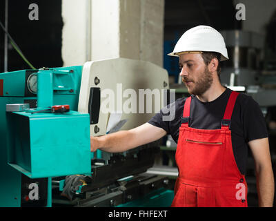 left side view of a worker, standing, wearing red overalls, and a white protective helmet, operating the control panel of an ind Stock Photo