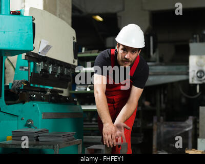 front view of a worker, wearing red overalls, and a white protective helmet, bending over to pick a bent piece of metal, next to Stock Photo