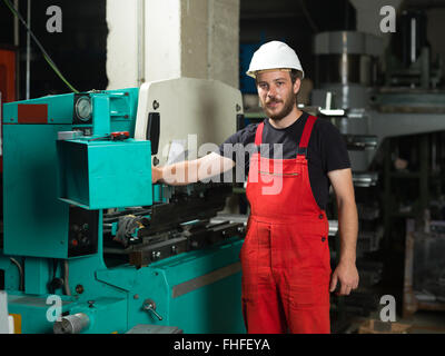 front view of a worker, standing, looking proud, wearing red overalls, and a white protective helmet, operating the control pane Stock Photo