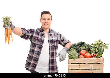 Young farmer holding a bunch of carrots and leaning on a crate full of vegetables isolated on white background Stock Photo