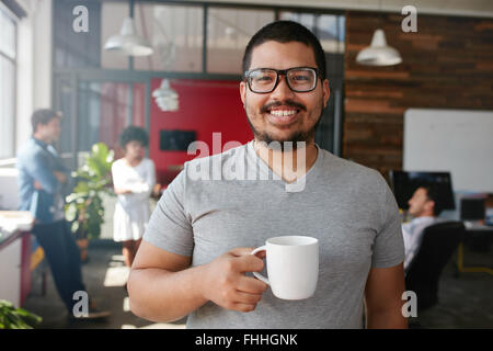 Portrait of smiling office worker having a coffee with his colleagues talking in the background. Stock Photo
