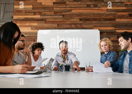 Portrait of team of young people having a meeting in office. Young men and women sitting around conference table during meeting. Stock Photo