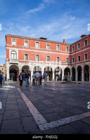 Colonnaded red stucco building around the sunlit patterned courtyard of Campo di San Giacomo di Rialto, Venice, Italy busy with tourists and locals Stock Photo