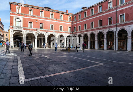 Colonnaded red stucco building around the sunlit patterned courtyard of Campo di San Giacomo di Rialto, Venice, Italy busy with tourists and locals Stock Photo