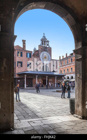 Campo and Chiesa di San Giacomo di Rialto with its large dialled clock, Venice, Italy, busy with people seen through an arch against a clear blue sky Stock Photo