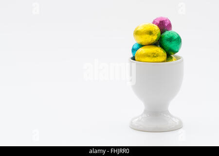 A white egg cup filled with small, chocolate Easter eggs on an isolated white background. Stock Photo