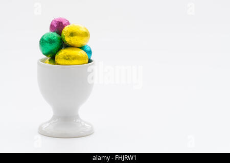A white egg cup filled with small, chocolate Easter eggs on an isolated white background. Stock Photo