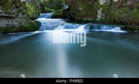 A small river between rocks at the riverside flows into a blue lagoon. Scene from a german forest. Deep clear water. Stock Photo