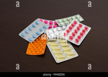 Heap of packs of pills and capsules (white, blue, green, yellow, orange, pink, red, wine-colored) on brown wooden background Stock Photo