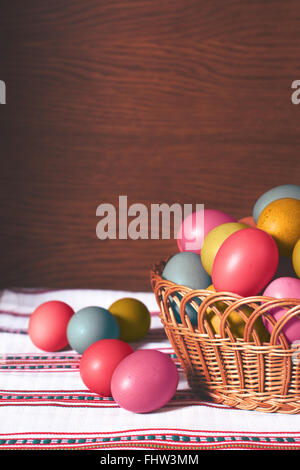 Vertical oriented image of basket full of colourful pastel easter eggs. On wooden background and white ornamented towel. Stock Photo