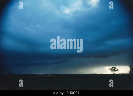 Heavy shower approaching in a dense, grey-blue cloud formation over countryside, with lone tree in the distance. Stock Photo