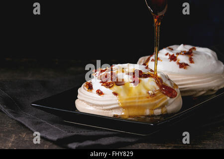 Black plate with Meringue dessert Pavlova with caramel powering by caramel sauce. On gray textile napkin over dark table. Stock Photo