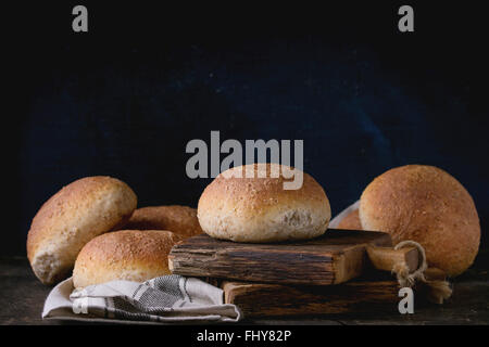 Fresh baked wholegrain buns on gray kitchen towel over small wooden chopping board with black background. Stock Photo